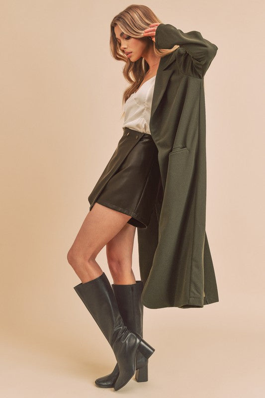 Woman wearing a long green coat, black skirt, and black knee-high boots on a beige background