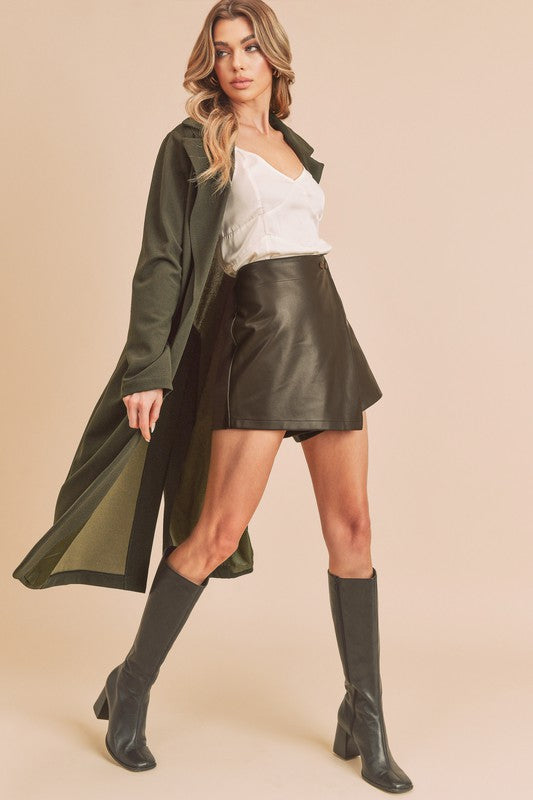 Woman wearing a green trench coat, white top, black leather skirt, and black boots on a beige background