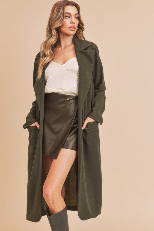 Woman wearing a long green coat over a white top and black leather skirt on a beige background