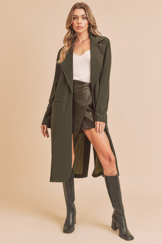 Woman wearing a long green coat, white top, black leather skirt, and black knee-high boots on a beige background