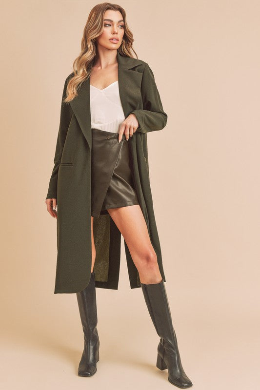 Woman wearing a long olive green coat, white top, and black boots on a beige background
